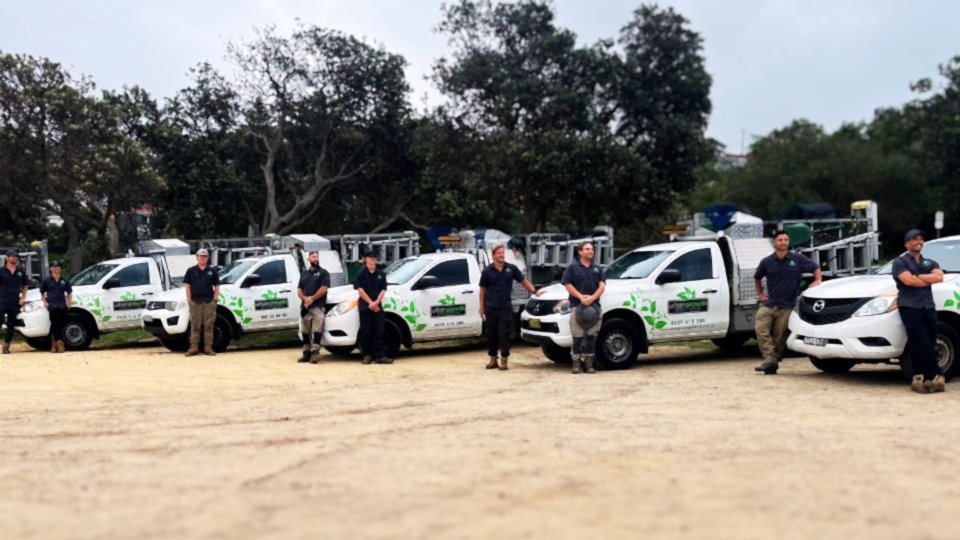 A dedicated gardening team stands ready with tools and smiles, beach backdrop on Sydney's Northern Beaches, NSW